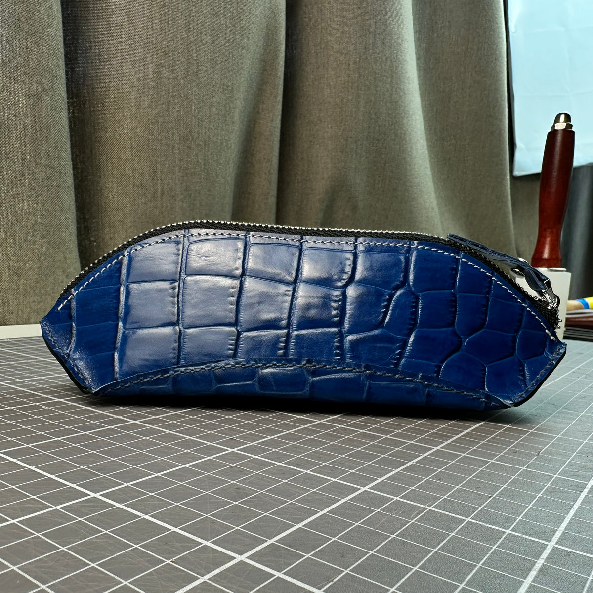Side view of a handcrafted blue alligator-embossed leather pencil case with white contrast stitching on a cutting mat.