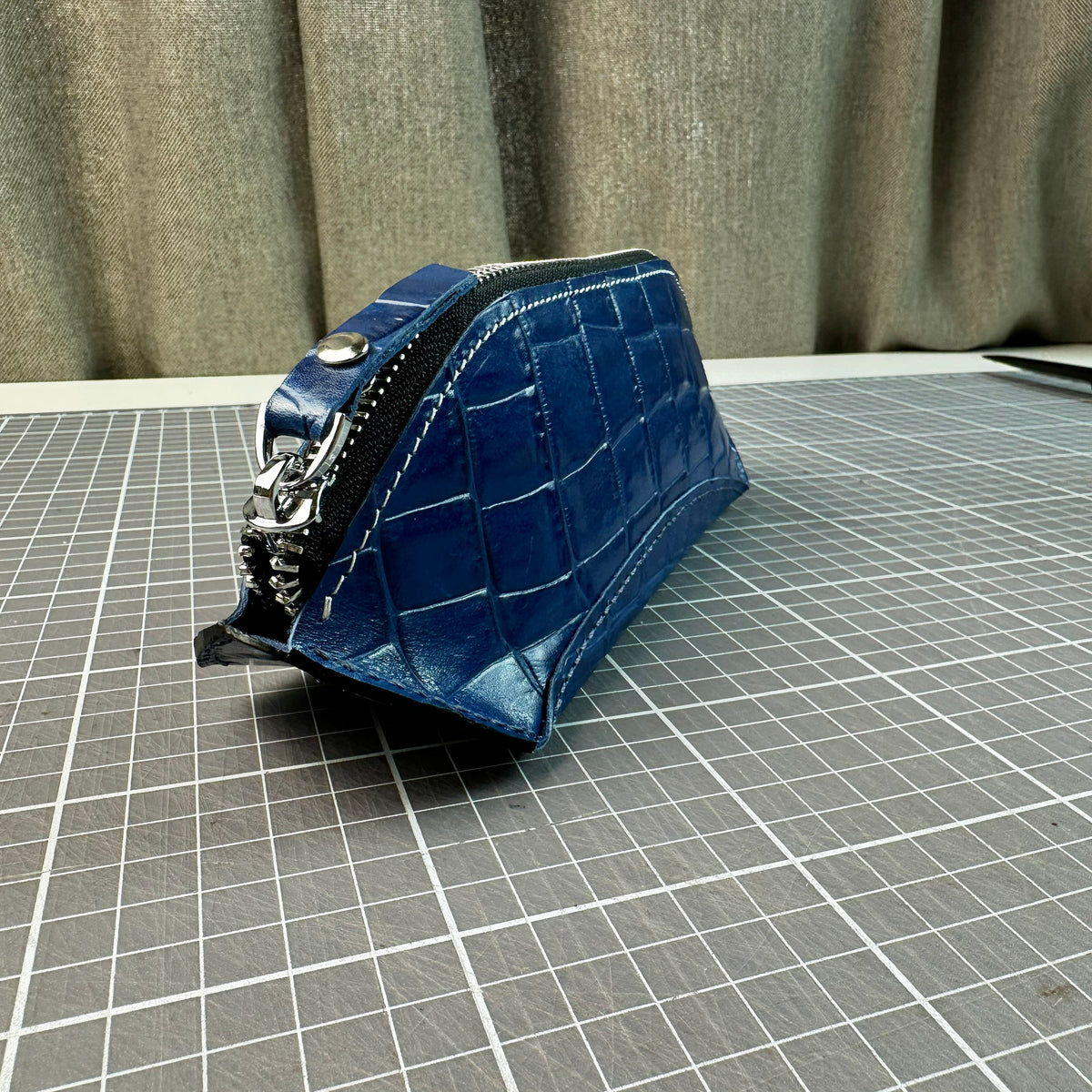 Blue crocodile-textured leather pencil case with metal zipper on a cutting mat showing a side profile view.