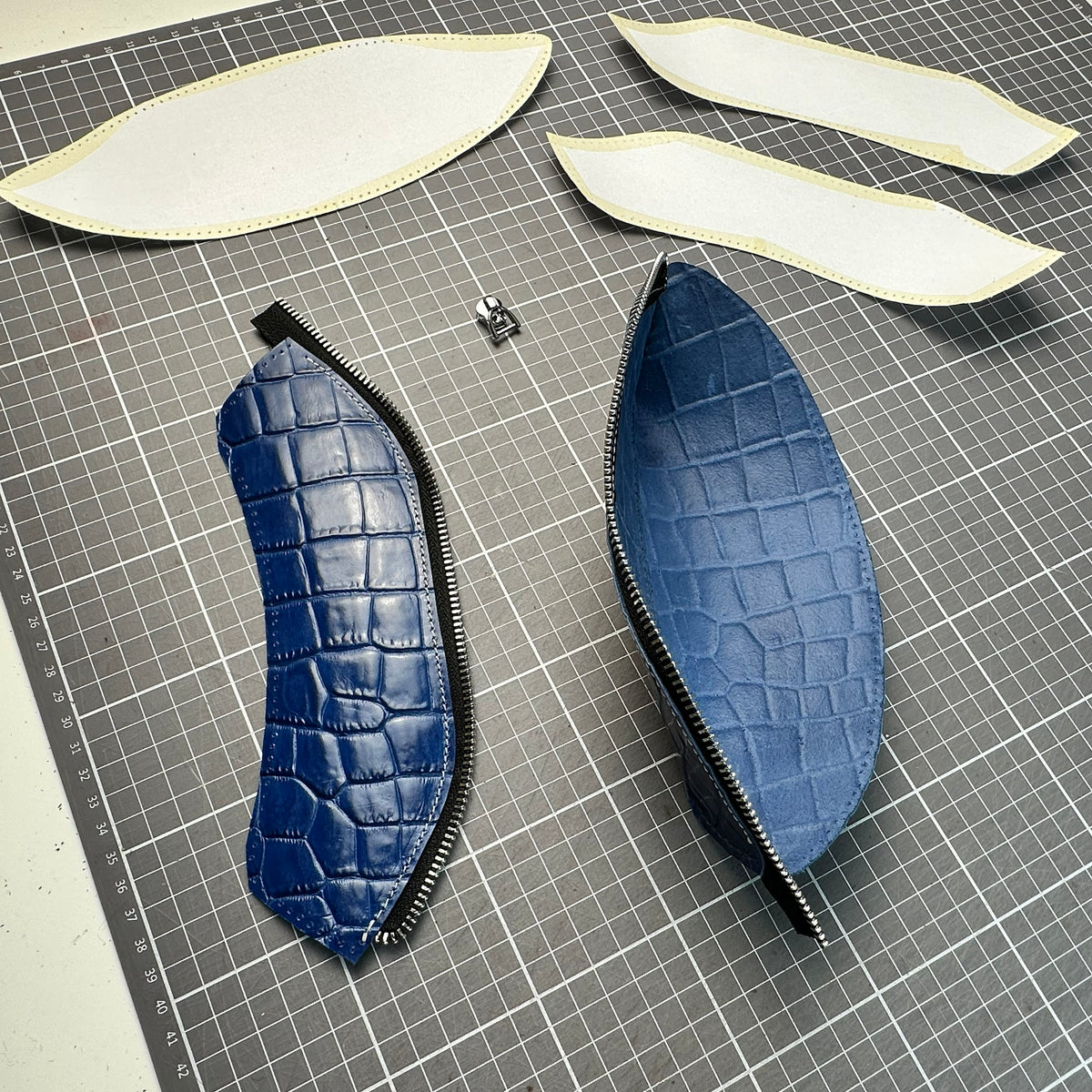 Blue faux-croc leather pencil case pieces with zippers laid out on a cutting mat with paper sewing templates.