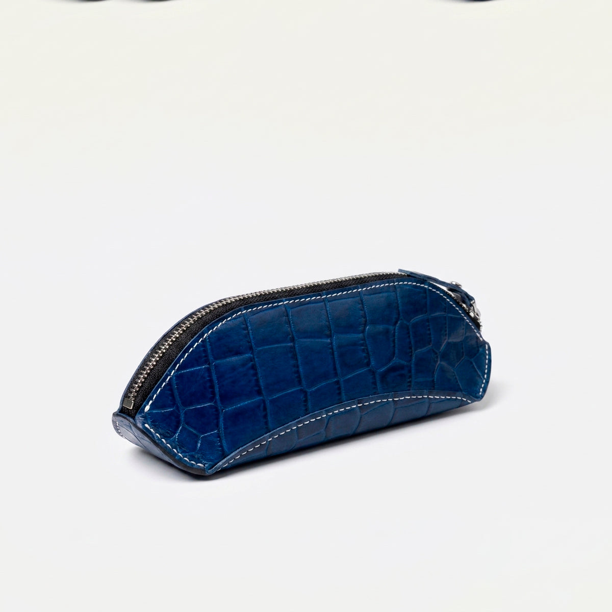 Blue crocodile-embossed leather pencil case with white saddle stitching and a silver zipper, angled side view.