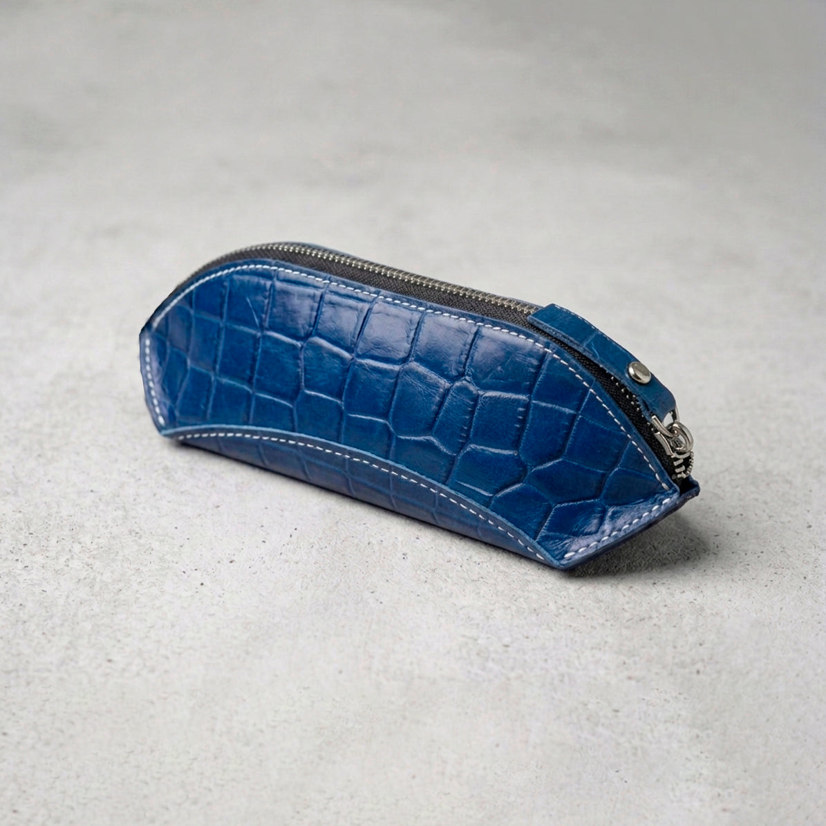 Blue crocodile-embossed leather pencil case with a silver zipper, captured from a high-angle side view.