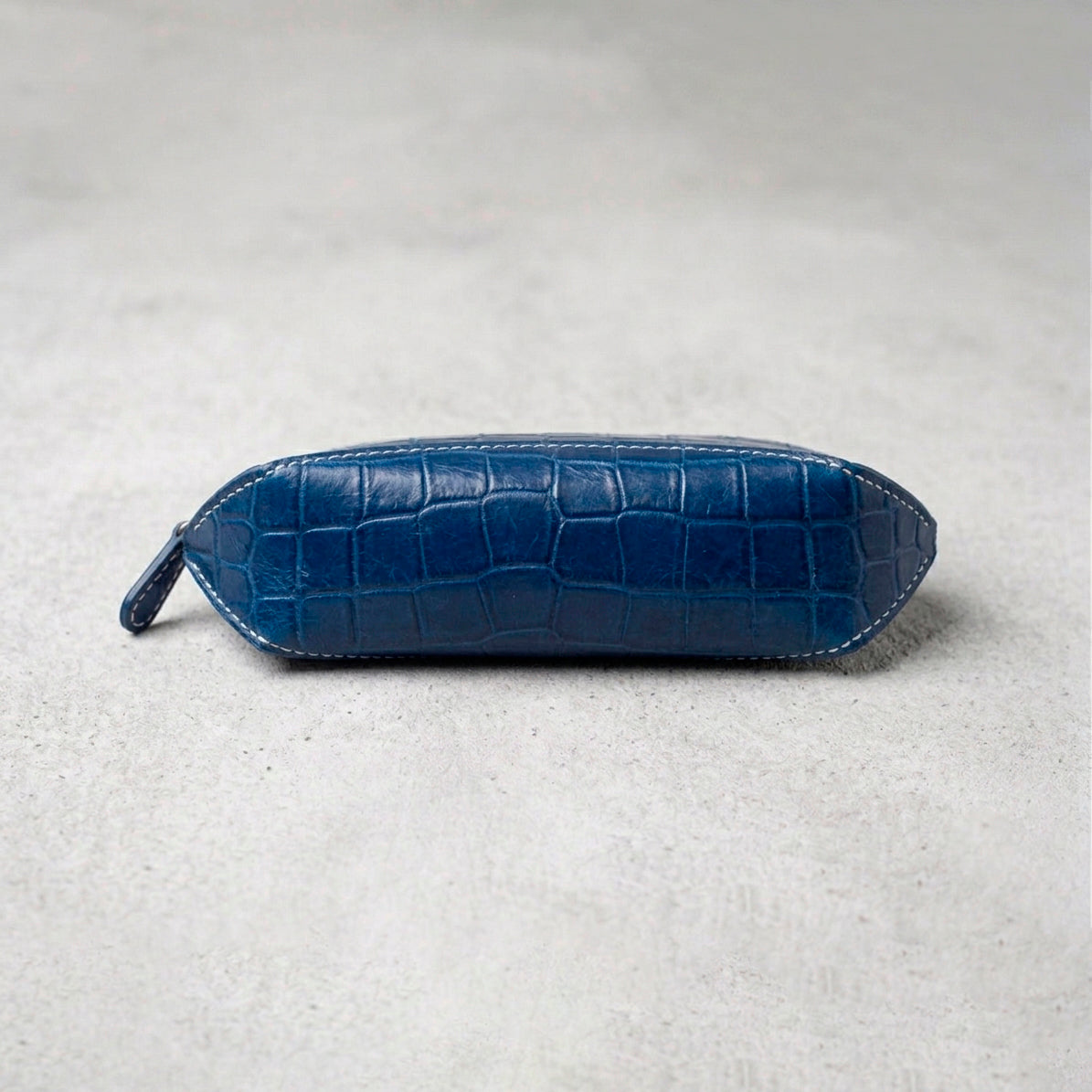 Side view of a blue crocodile-embossed leather pencil case with white contrast stitching on a grey concrete surface.