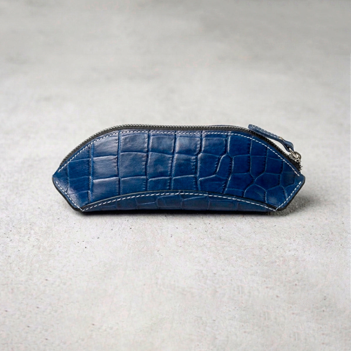Side view of a blue crocodile-embossed leather pencil case with a silver zipper and white contrast stitching.