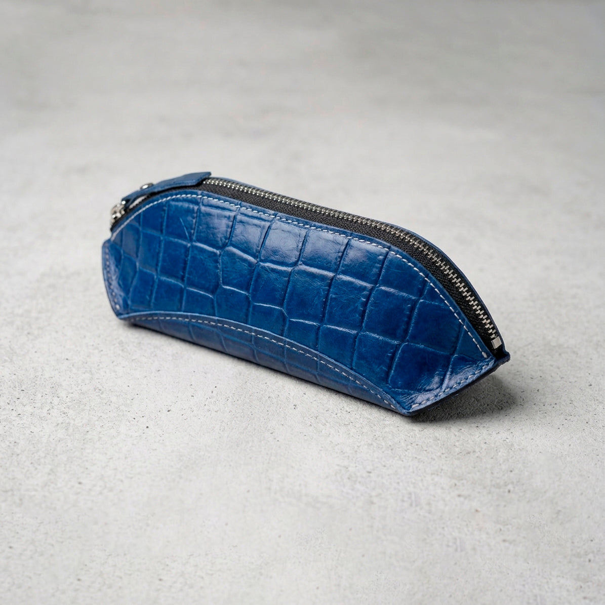 Blue crocodile-embossed leather pencil case with top zipper, viewed from a high angle on a light gray surface.