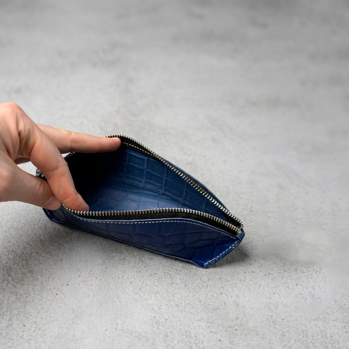 Hand opening a blue crocodile-embossed leather pencil case, showing the spacious interior and polished metal zipper.