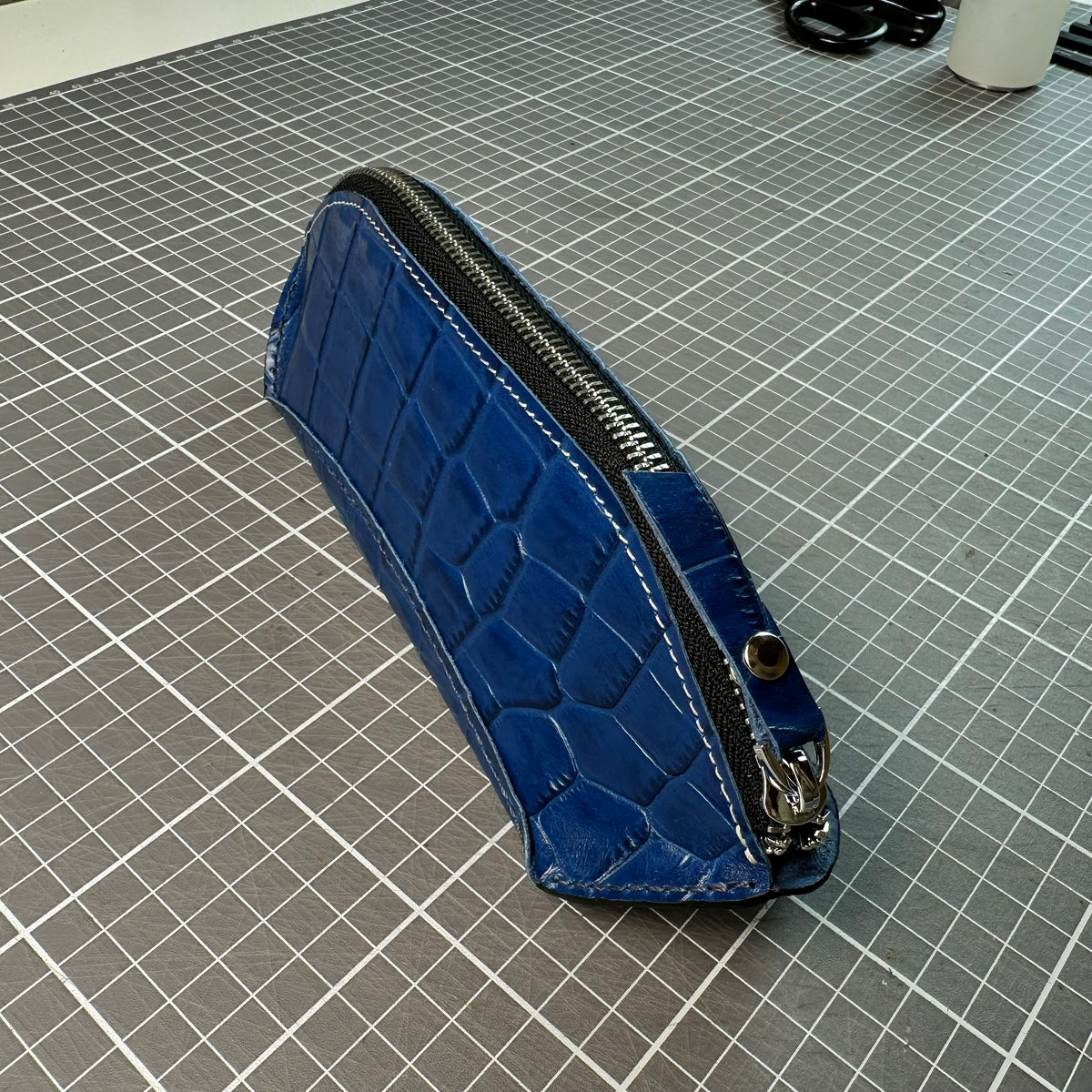 Blue embossed leather pencil case with a metal zipper, angled view showing side stitching on a cutting mat.